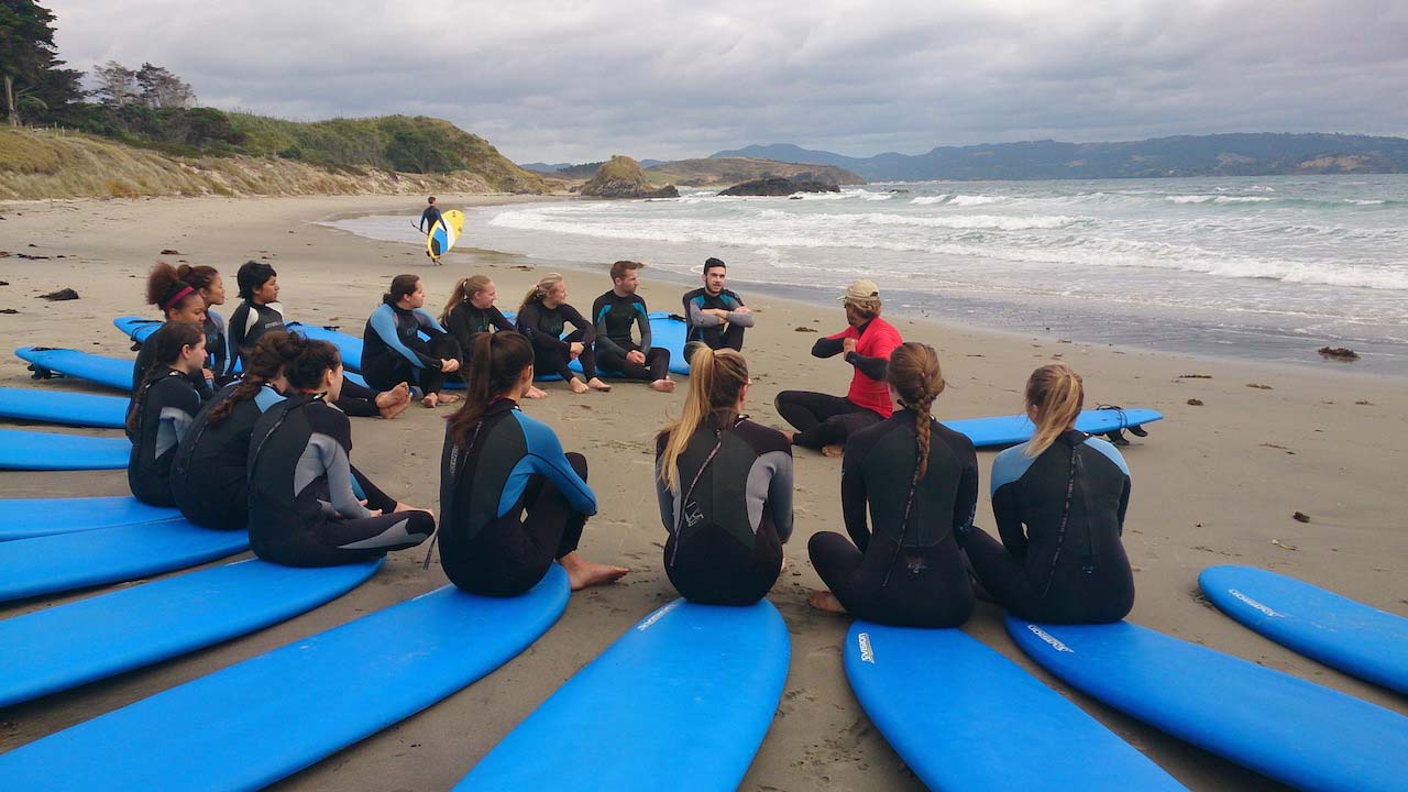 A group of students dressed in wetsuits sit in a circle on their surfboards on the beach listening to the instructor