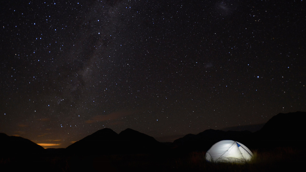 Stars shine bright across the dark sky and a tent stands illuminated in Arthur's Pass National Park, New Zealand