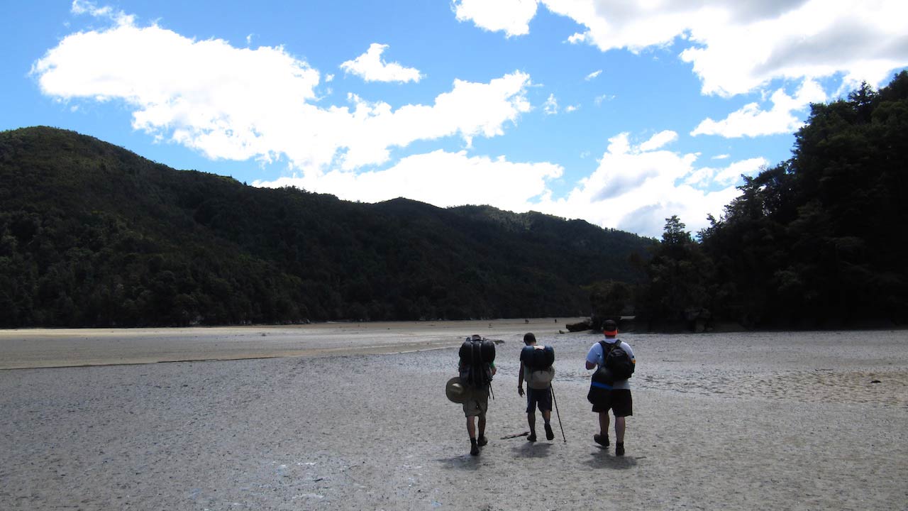 Three people walk with backpacks on flat ground towards the mountains near Wellington, New Zealand