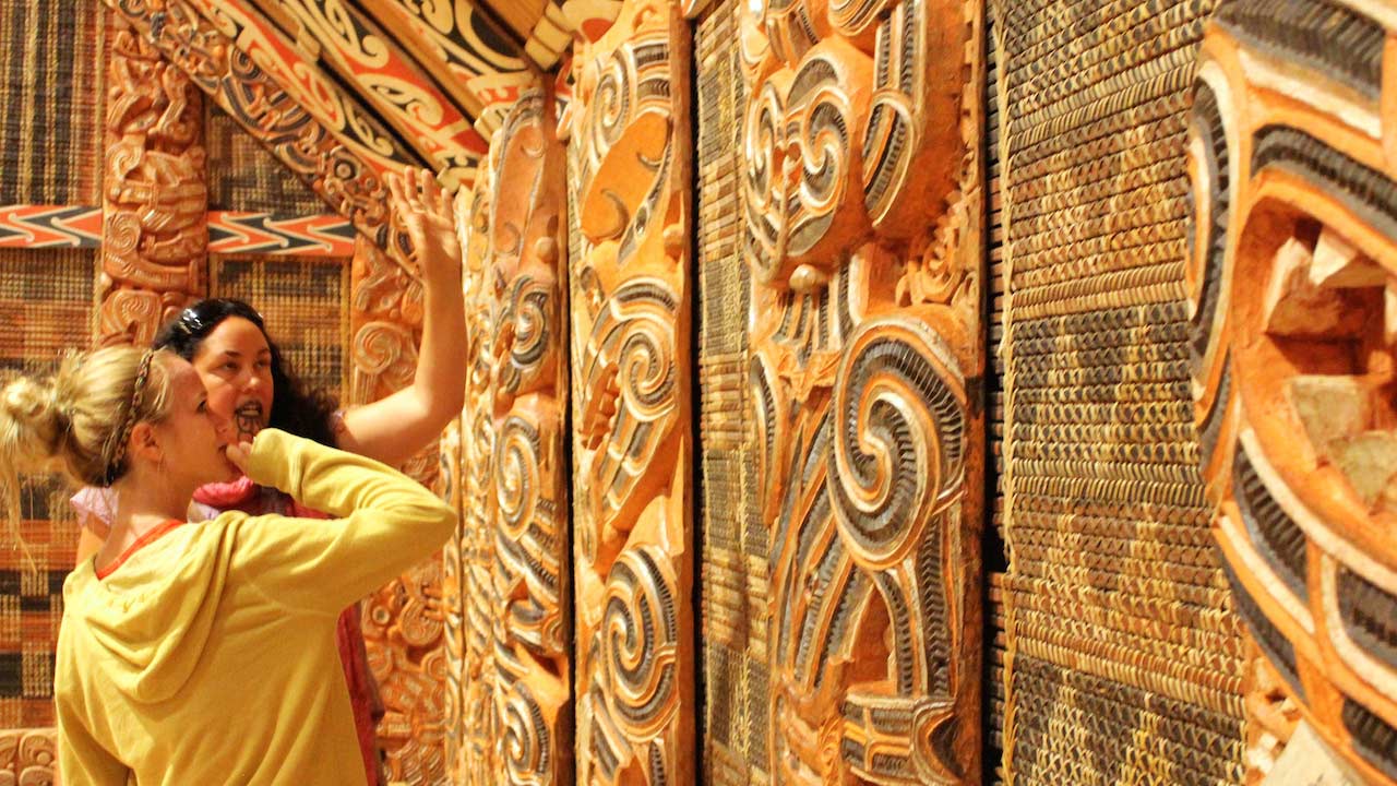 Two women discuss a wall of traditional Maori carvings