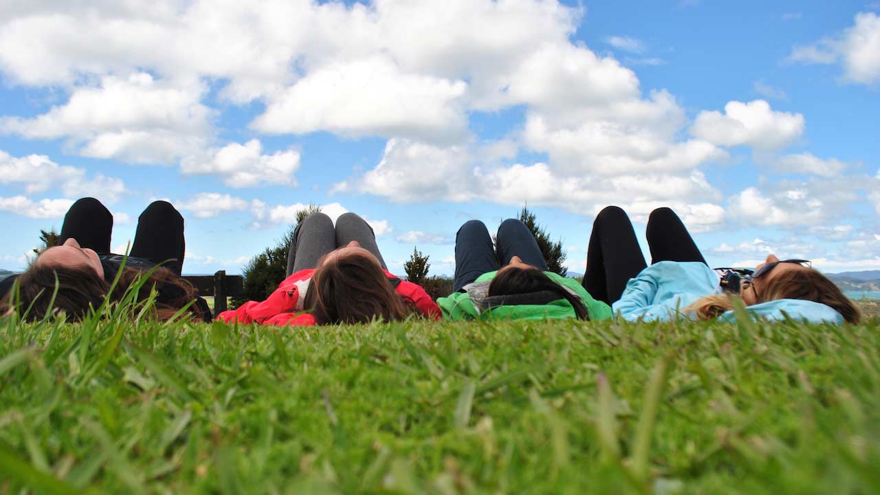 Four students lay in the grass and look up at a blue sky