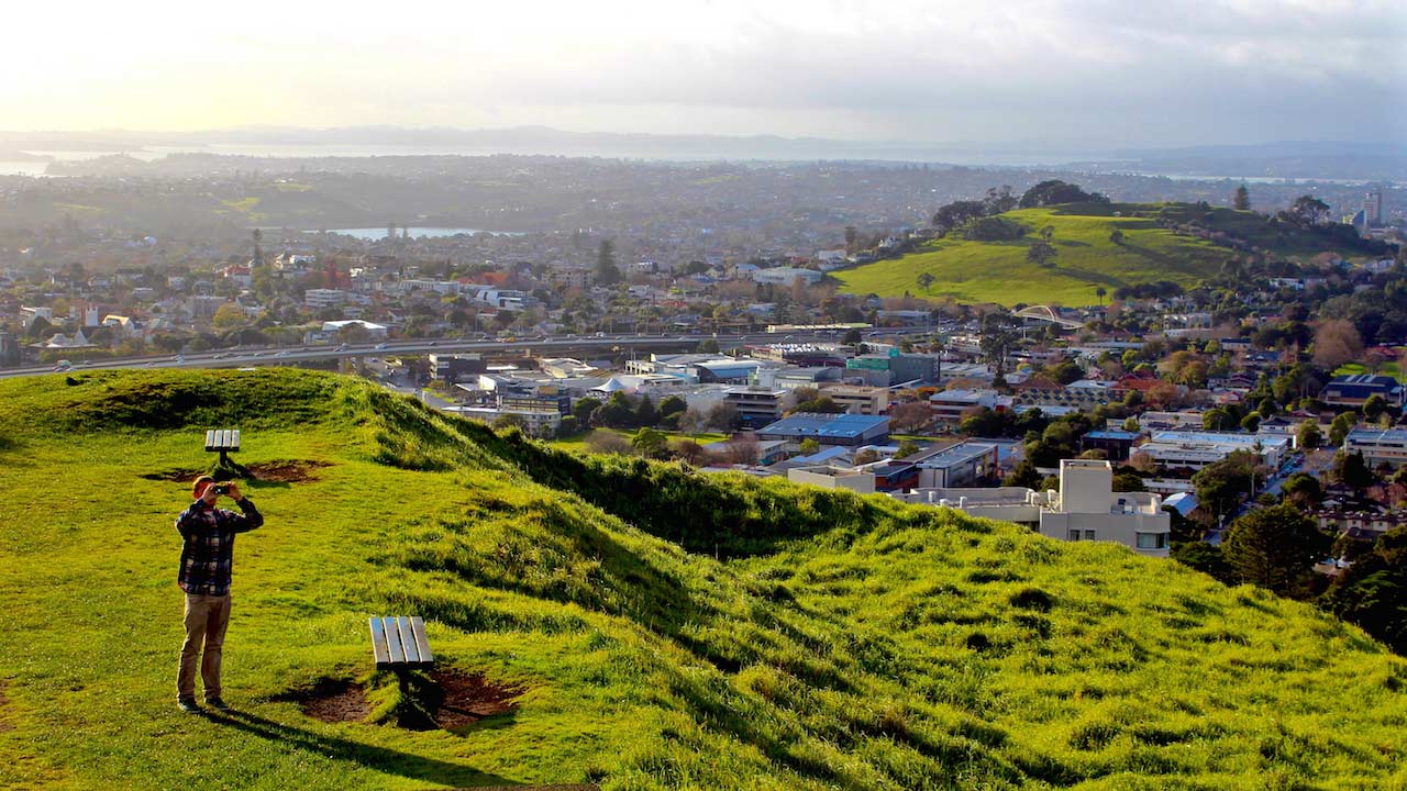 A man stands on a grassy hill overlooking Auckland and looks at the sky with binoculars
