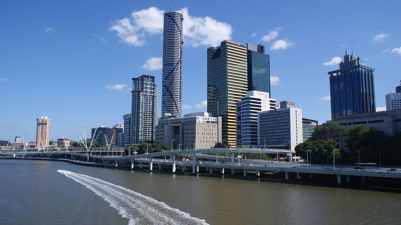 A boat cruises down the Brisbane River past the city's business district on a clear, sunny day