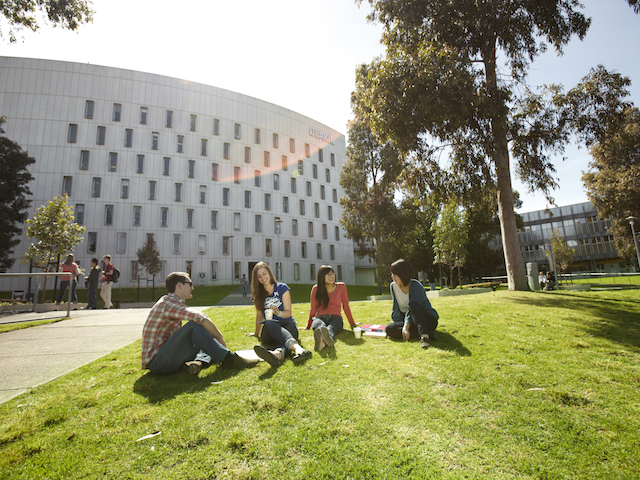 Four students sit on a patch of grass in front of a building on Deakin's campus