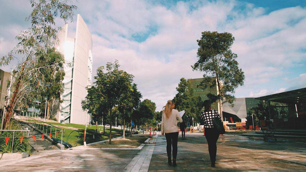 Two women walk along a pathway between buildings on Deakin's campus on a cloudy day