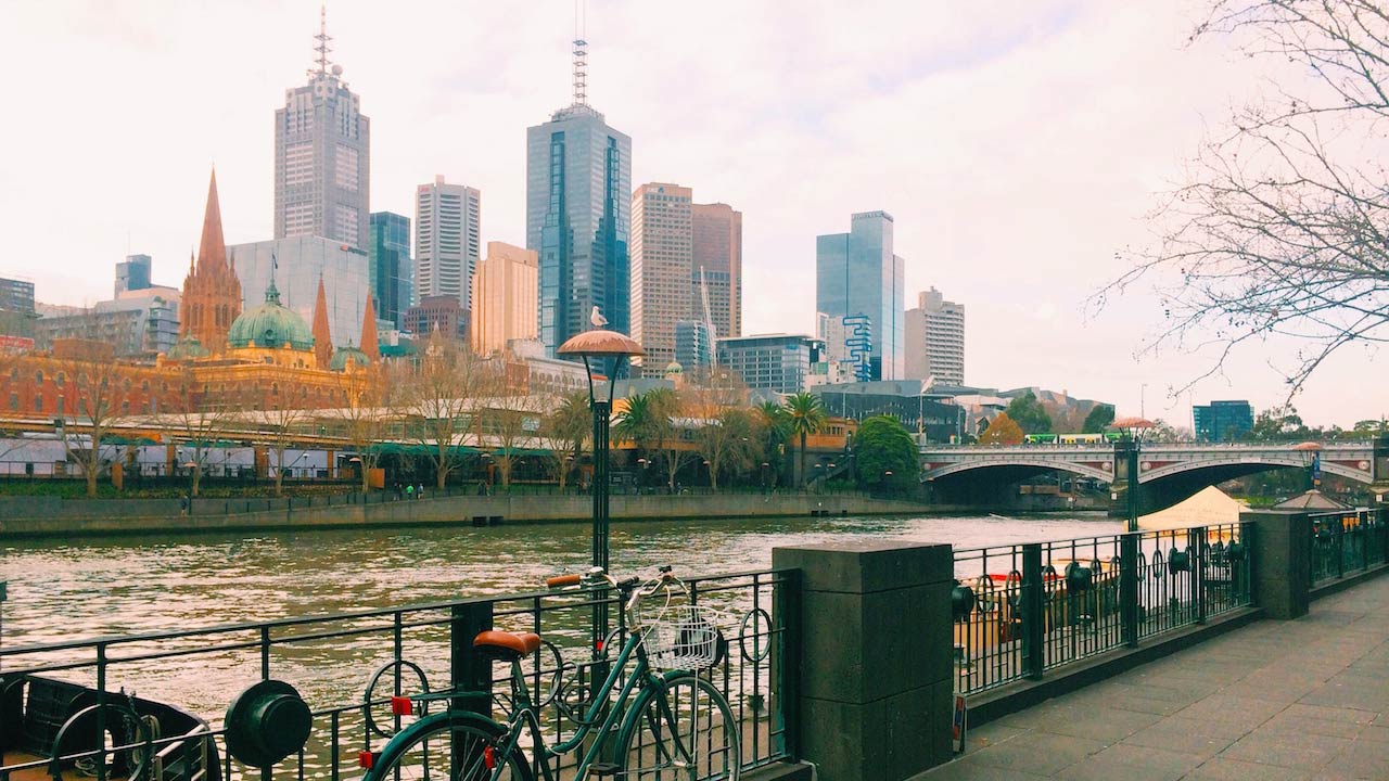 A bicycle leans against an iron fence in front of a river in Melbourne with the city's skyline as a backdrop