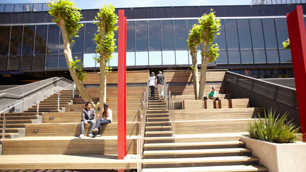 A few people sit on large steps outside of a building on Deakin's campus