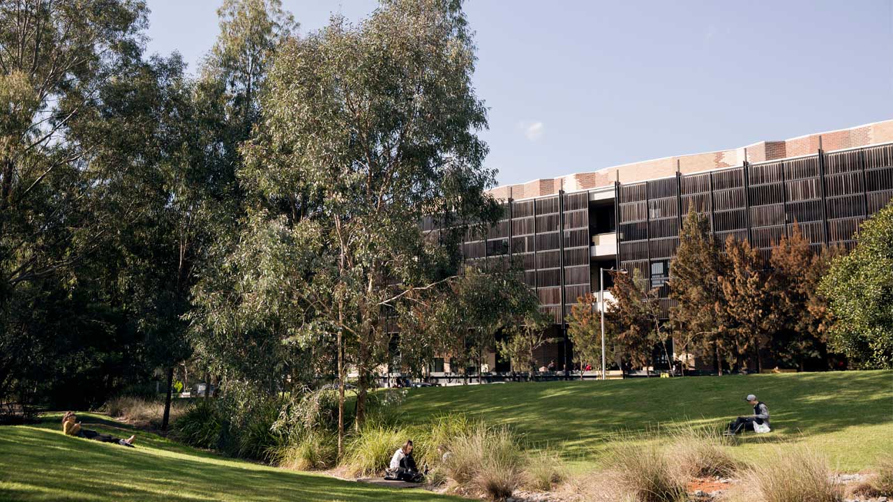 Three students scattered across a grassy quad on Deakin's campus study and relax