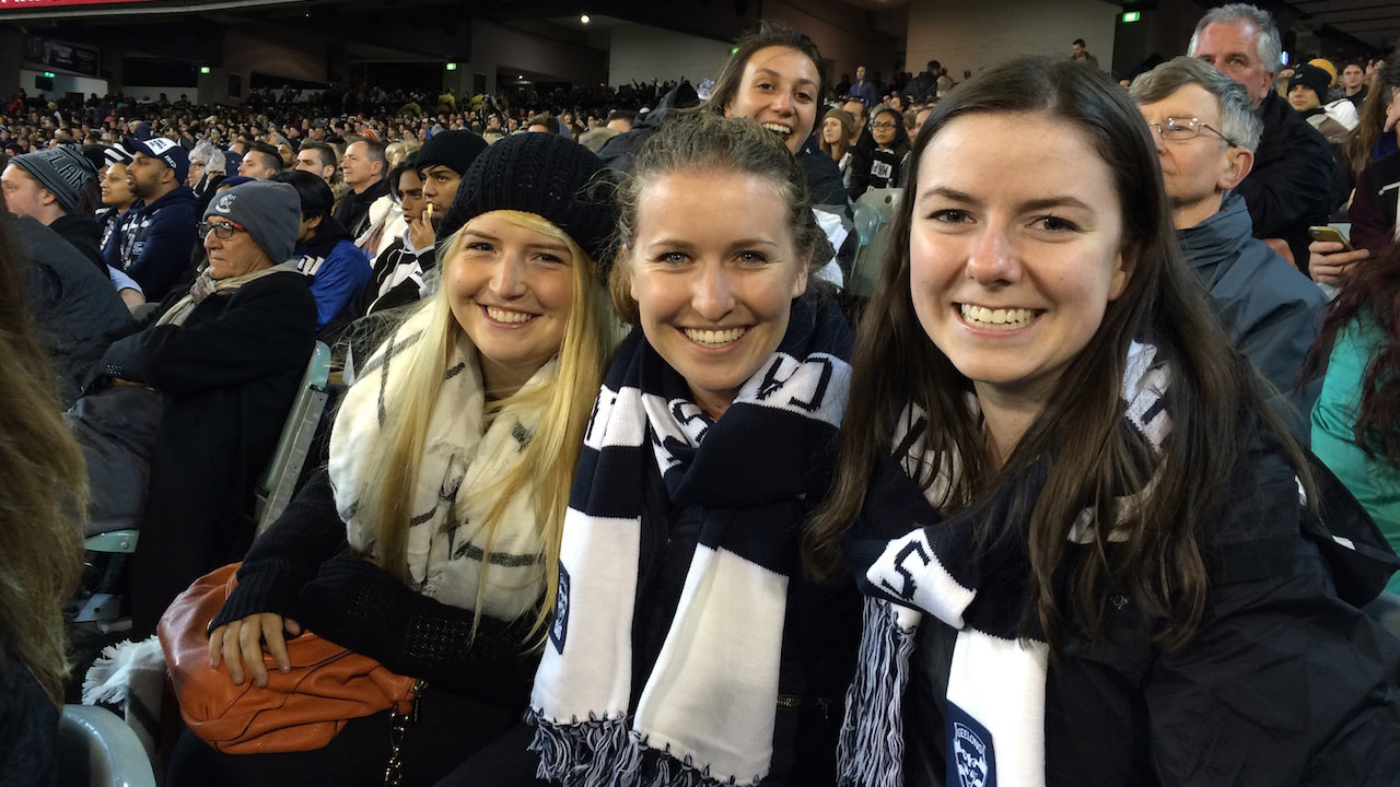 Four women dressed in spirited clothes sit smiling in a crowded stadium