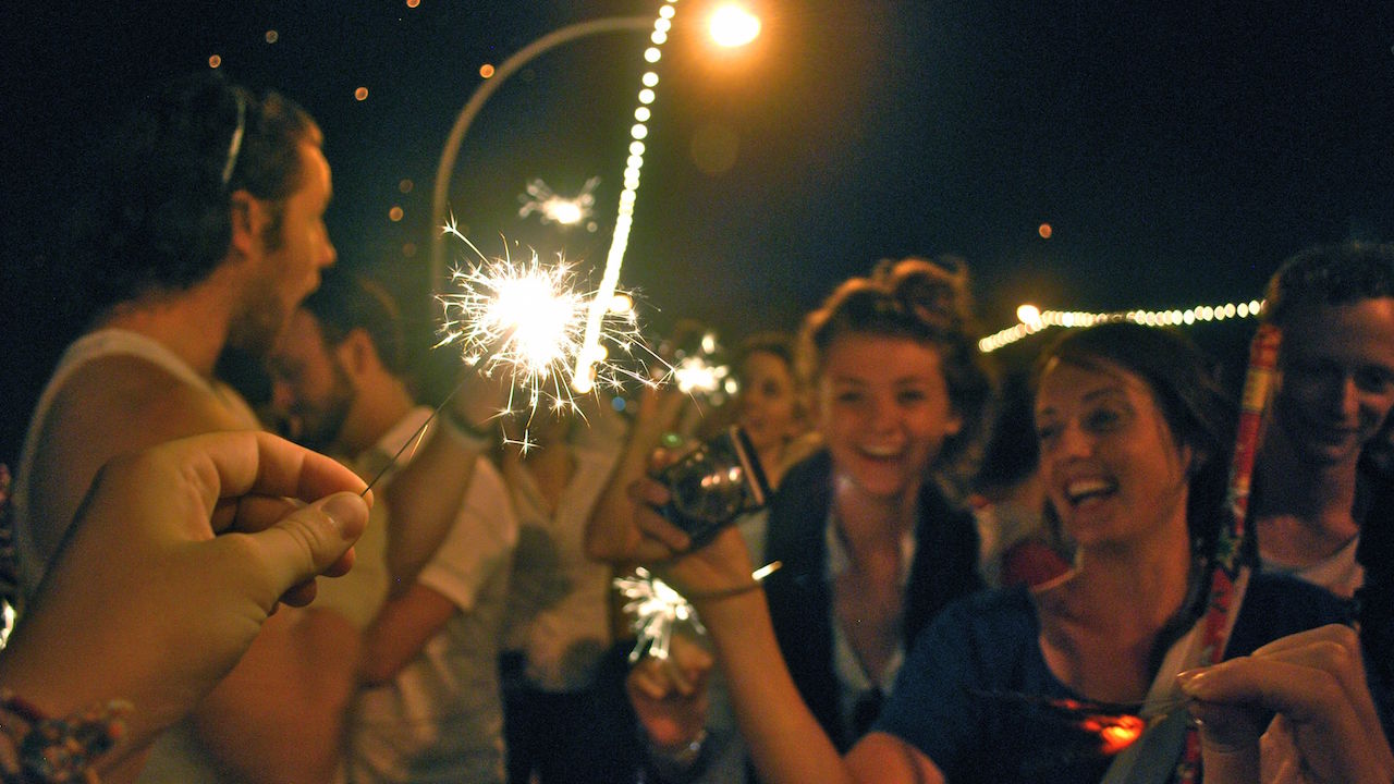 A hand holding a lit sparkler in front of girls' excited faces at nighttime in Thailand