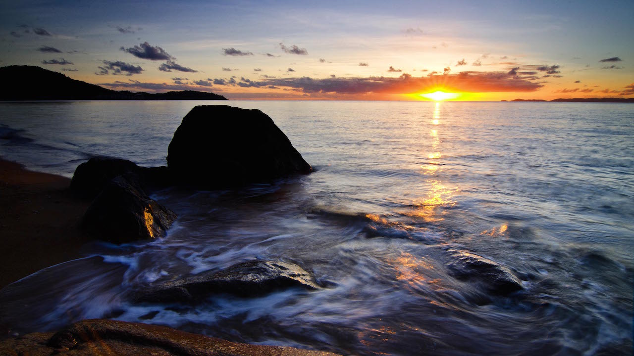 The sun sets behind the horizon as waves crash against a rock on the shore of Magnetic Island in Australia