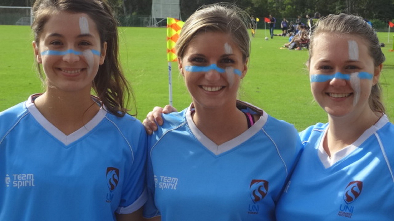 Three women with blue and white paint on their faces show their school spirit on the stadium's field on University of Newcastle's campus