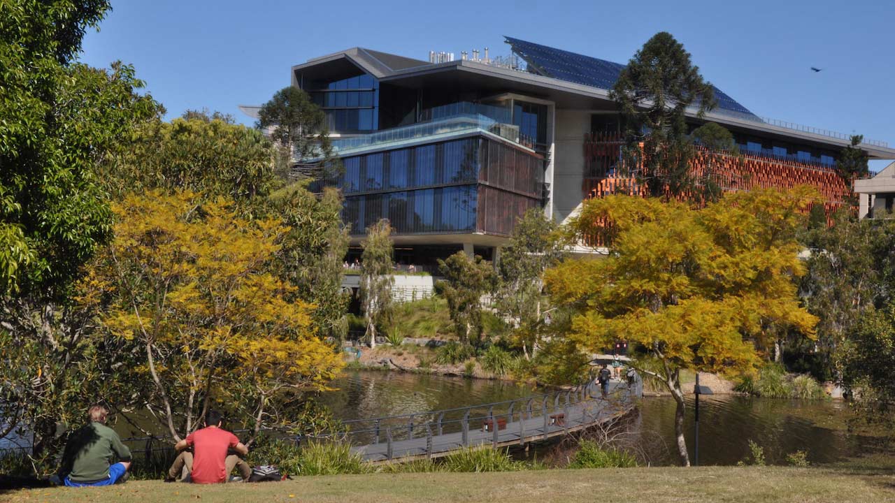 Students sit with their backs to the camera, facing the lake, walkway and building on UQ's campus