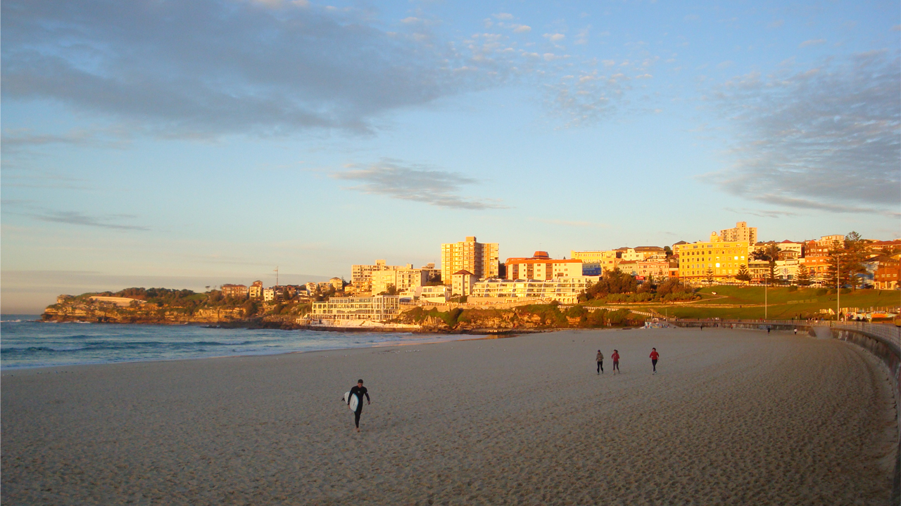 A handful of people walk along the beach as the sun illuminates a nearby neighborhood in Sydney