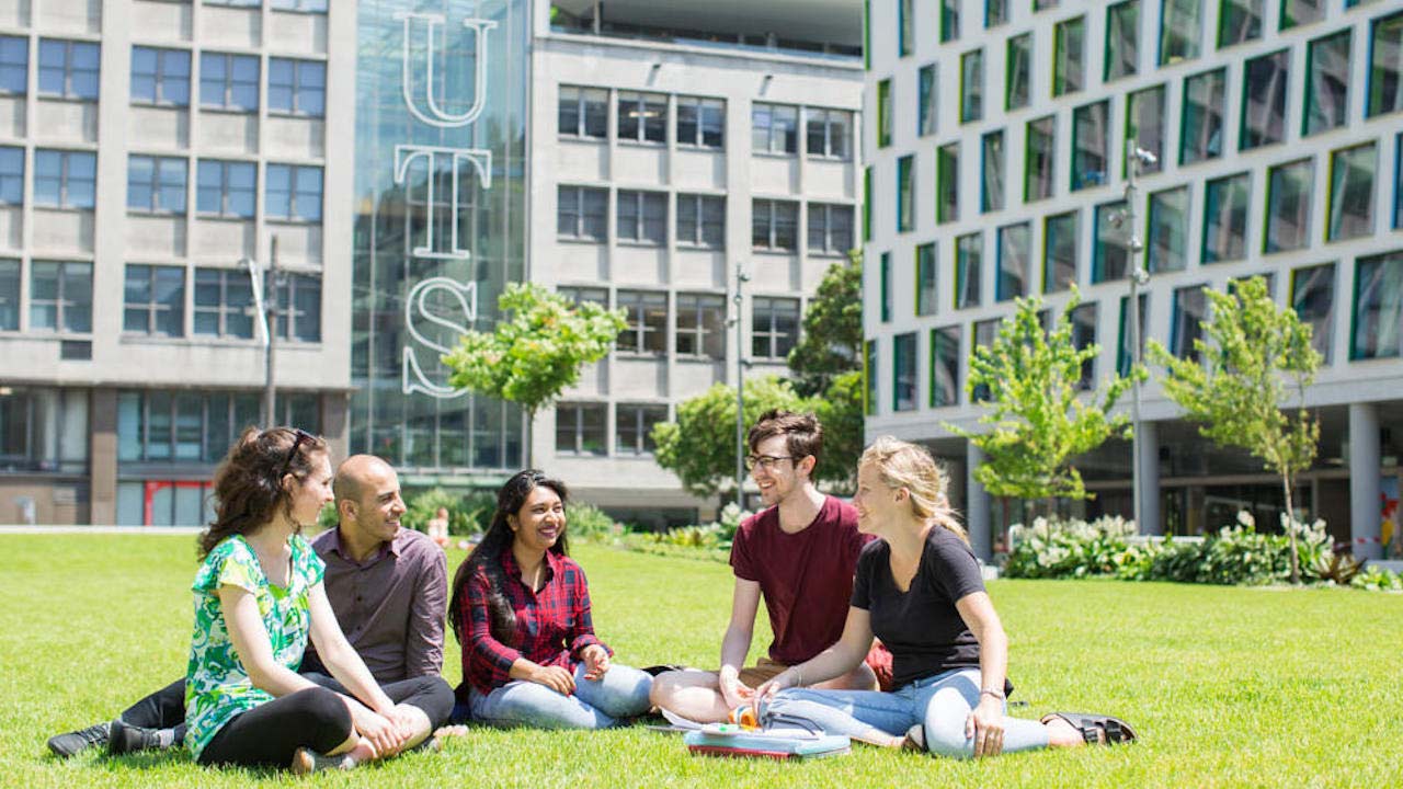 A group of people sit chatting on a grassy quad surrounded by buildings on UTS's campus