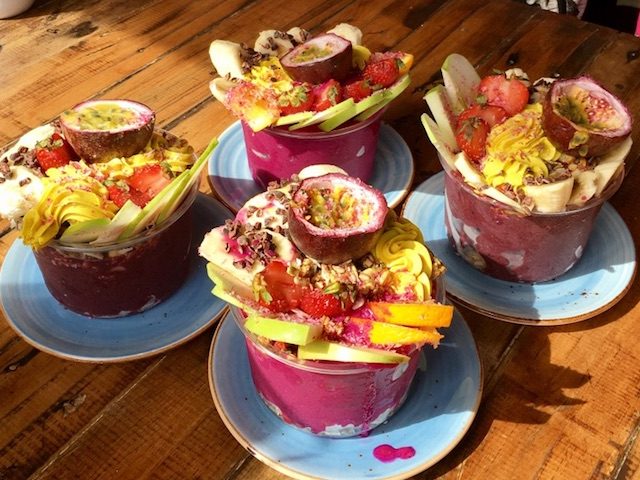 Four Acai bowls topped with granola and fresh fruit sit on a wooden table