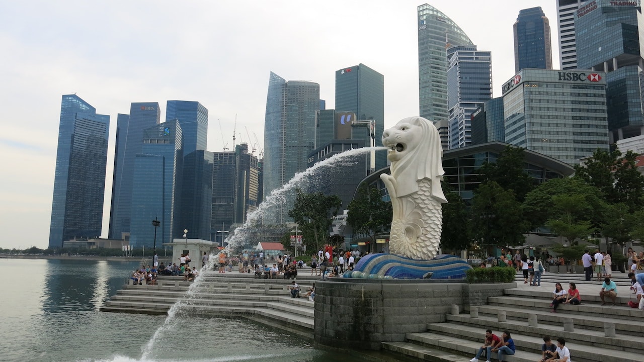A fountain sprays water into the harbor as people sit on stairs in front of the Singapore skyline